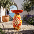 Red Mediterranean-style glass bird bath with a vibrant red, orange, green, and cream patterned pedestal, displayed on a stone patio surrounded by olive trees and lavender.