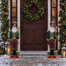 Two decorative Santa figures in matching green, burgundy, and gold outfits displayed on either side of a front door, surrounded by illuminated holiday garland, wreaths, lanterns, and snow. Each Santa stands on an ornate red and green base.