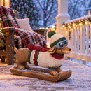 Winter dachshund statue wearing a knit hat, goggles, sweater, and scarf displayed on a snow-covered porch at night with string lights.