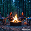 Black circular metal fire pit with burning logs and flames, surrounded by four people sitting in camping chairs in a wooded forest. Image labeled “AI Generated Performer.”