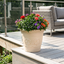 Cream-colored decorative planter filled with vibrant red, purple, pink, and orange flowers and lush green foliage, displayed on a modern outdoor deck with patio seating.