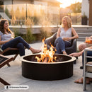 Black circular metal fire pit with burning logs and flames, surrounded by three people relaxing on a patio at sunset. Image labeled “AI Generated Performer.”