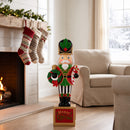Christmas nutcracker decoration dressed in a festive red, green, black, and gold uniform, holding a holiday wreath and candy cane while standing on a Merry Christmas base. Displayed on a wooden floor beside a cozy fireplace decorated with Christmas stockings and garland, with beige furniture and snowy windows in the background.