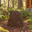 Dark brown decorative faux rock cover with a tall, tapered shape and realistic stone texture, placed in a mulch-covered forest landscape surrounded by ferns, trees, and a rustic log cabin.