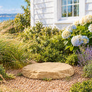 Tan decorative faux rock cover with a flat, natural stone-textured surface and irregular edges, placed in a gravel-covered garden bed surrounded by ornamental grasses, flowering hydrangeas, lavender, and shrubs beside a white house.