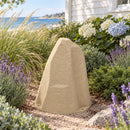 Beige faux rock utility cover in a coastal garden beside a white shingle-sided home, surrounded by ornamental grasses, lavender, hydrangeas, evergreen shrubs, and gravel, with the ocean visible in the background.