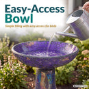 Crackled Glass Mosaic Bird Bath in iridescent purple shown outdoors in a garden as water is poured into the wide, shallow bowl, highlighting easy filling and access for birds.