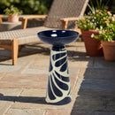 Navy blue and cream decorative pedestal bird bath displayed on a stone patio, surrounded by outdoor furniture, greenery, and potted flowers.