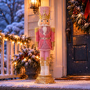 Pink, white, and gold Christmas nutcracker displayed outdoors in a snowy, decorated entryway, surrounded by evergreen garland, warm holiday lights, and a dark front door.