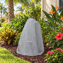 Light gray faux rock utility cover with a tall, tapered shape and realistic stone texture, displayed in a tropical garden with palm trees, bird of paradise flowers, hibiscus blooms, and lush green foliage beside a stucco house.