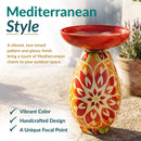 Mediterranean-style red glass bird bath with a vibrant red, orange, green, and cream patterned pedestal, displayed on a sunlit stone patio. Text highlights its vibrant color, handcrafted design, and unique focal point.