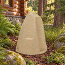 Beige faux rock utility cover placed in a mulched garden bed beside a rustic log cabin, surrounded by ferns, ornamental grasses, evergreen trees, and natural landscaping.