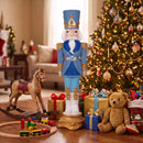 Blue, white, and gold Christmas nutcracker displayed indoors beside a decorated Christmas tree, wrapped gifts, a rocking horse, toys, and a teddy bear.