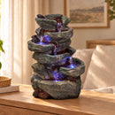 Tabletop cascading rock fountain with multiple tiers of textured gray stone basins, flowing water, and vibrant blue LED lights, displayed on a wooden console table in a cozy living room.