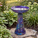 Crackled Glass Mosaic Bird Bath in iridescent purple shown in a landscaped garden, featuring a water-filled purple bowl and tapered pedestal with shimmering blue, purple, and multicolor iridescent mosaic accents.