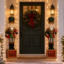 Two green and red Christmas nutcracker statues holding festive wreaths and standing on matching “Merry Christmas” bases, flanking a front door decorated with a large holiday wreath, lanterns, and illuminated string lights.