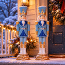 Two blue and gold nutcracker soldiers standing in the snow outside a warmly lit home, surrounded by festive Christmas trees and winter decorations. Both wear blue uniforms, white beards, and ornate gold-trimmed crowns.