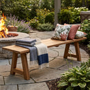 Sunnydaze Teak Trestle Bench in a backyard by a firepit, with cozy items on the top like blankets and pillows. 