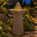 Outdoor pedestal fountain with a tiered pyramid water feature and illuminated water, surrounded by lush landscaping and a stone pathway at dusk.