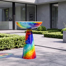 Colorful glass birdbath with vibrant red, blue, green, and yellow swirls displayed on a modern outdoor patio surrounded by manicured greenery.