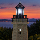 Weathered brown and green lighthouse statue with an illuminated lantern displayed at dusk overlooking a coastal landscape.