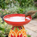 Close-up of a red Mediterranean-style glass bird bath with a vibrant red, orange, and green patterned pedestal, featuring a bird drinking from the water.