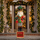 Green and red Christmas nutcracker statue holding a festive wreath and standing on a decorative “Merry Christmas” base in front of a warmly lit entryway decorated with illuminated holiday garland.