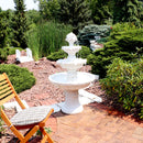 A white 3-tier outdoor fountain sits in a garden surrounded by trees and shrubs with water flowing down
