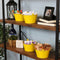 Four yellow galvanized steel buckets with handle filled with various items placed bookshelf containing books and pictures.
