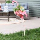 Outdoor flamingo garden statue with a planter full of flowers staked in the grass near an outdoor patio 