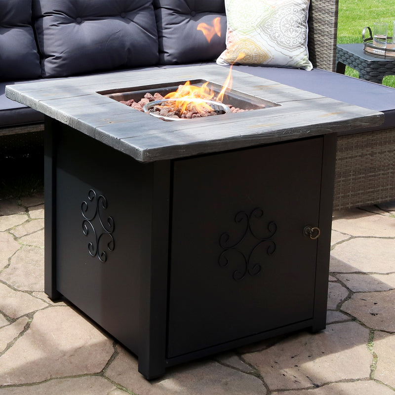 Square, black gas fire pit with gray faux wood top and fire burning in the center surrounded by lava rocks.