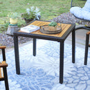 Square faux wicker patio table with steel frame and acacia wood surface sitting on outdoor patio with floral rug in between two patio chairs