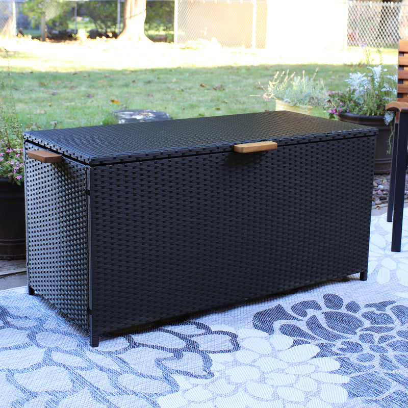 Black resin rattan deck box with acacia wood handles outdoors on a floral rug