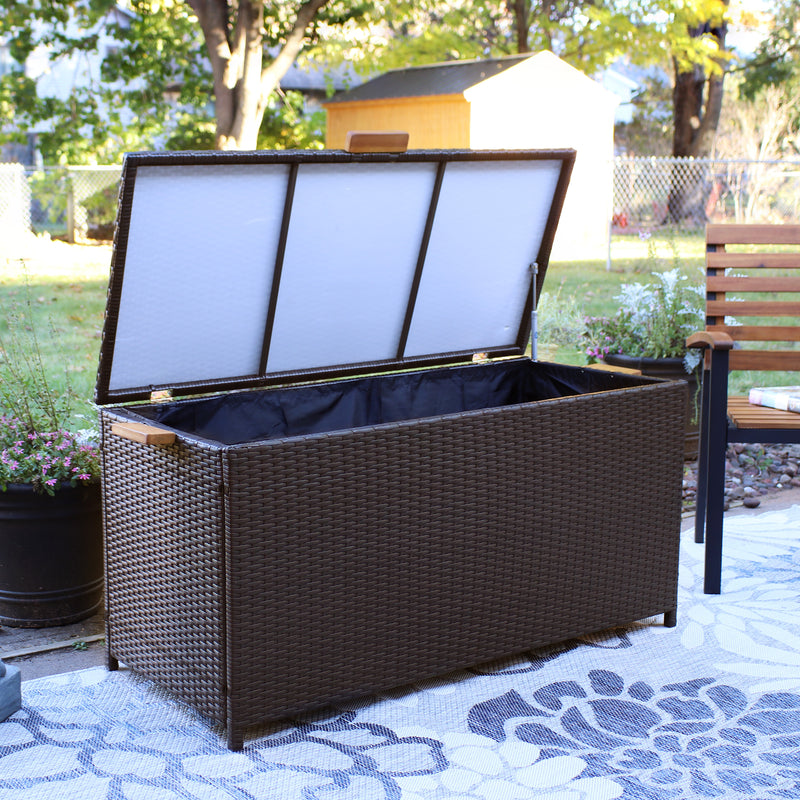 Brown resin rattan deck box and acacia wood handles with lid open outdoors on a floral rug