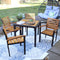 Square faux wicker patio table with steel frame and acacia wood surface sitting on outdoor patio with floral rug surrounded by four patio chairs